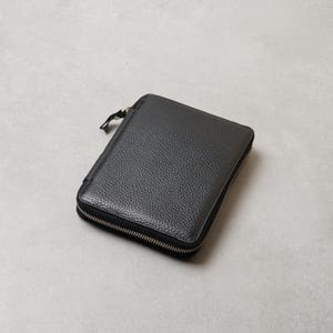 May include: A rectangular black wallet with a textured surface and a silver-toned metal zipper that runs along three sides. The wallet features a small, matching black zipper pull tab attached to the main zipper slider on the top left corner.