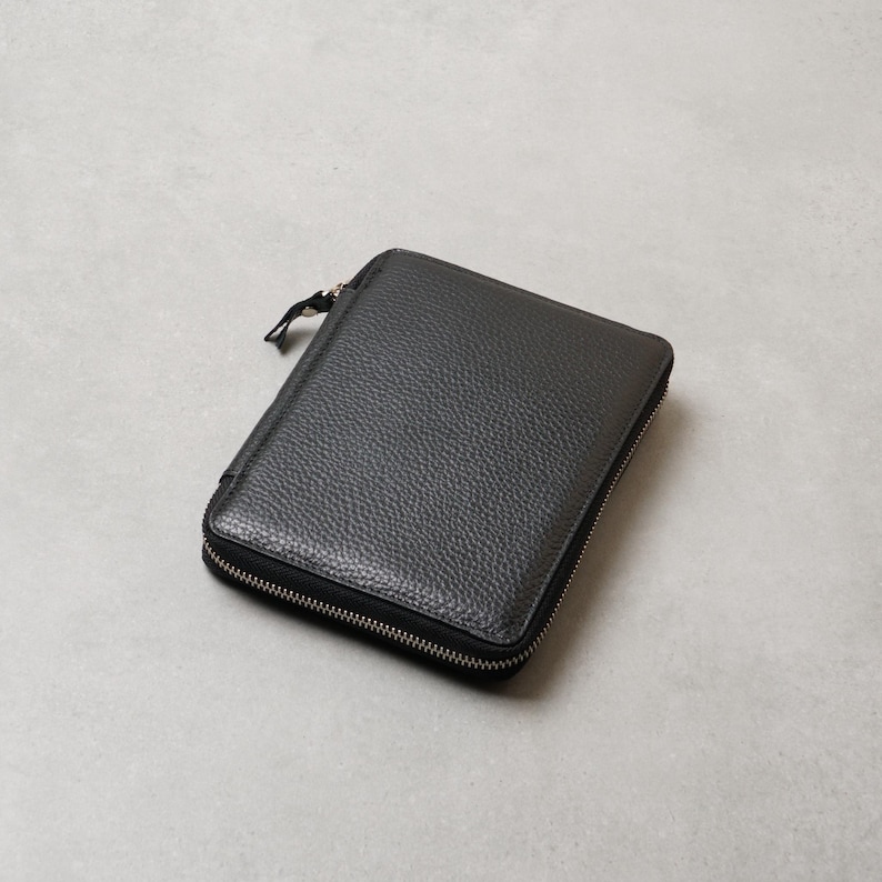 May include: A rectangular black wallet with a textured surface and a silver-toned metal zipper that runs along three sides. The wallet features a small, matching black zipper pull tab attached to the main zipper slider on the top left corner.