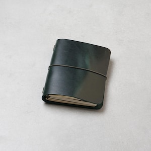 Peut inclure: Un carnet en cuir vert foncé avec une finition mate et lisse, maintenu par un élastique fin et sombre enroulé horizontalement autour du centre. Le carnet présente des pages de couleur crème visibles à l'intérieur de la couverture.