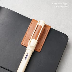 May include: A tan leather pen holder attached to a black notebook cover. The holder features visible stitching and secures a cream-colored fountain pen with a tan barrel accent. Text on the image reads: Can hold 1 regular fountain pen...