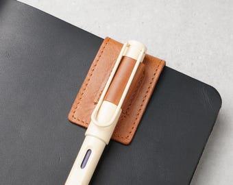 May include: A tan leather pen holder attached to a black notebook cover. The holder features visible stitching and secures a cream-colored fountain pen with a tan barrel accent. Text on the image reads: Can hold 1 regular fountain pen...
