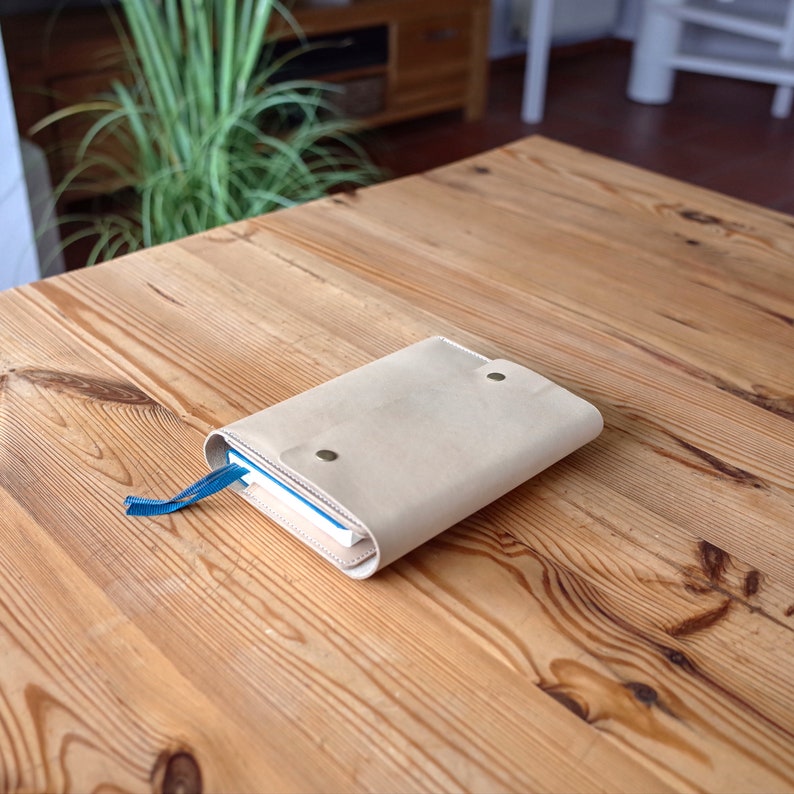 May include: A beige leather notebook cover with a blue ribbon bookmark. The cover has two metal snaps and is lying on a wooden table.