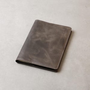 Peut inclure: Un journal en cuir gris avec un aspect us&eacute; et textur&eacute;. La couverture est d'une nuance de gris sourd, avec des zones plus sombres cr&eacute;ant un effet mouchet&eacute;. Le journal est rectangulaire et semble ferm&eacute;.