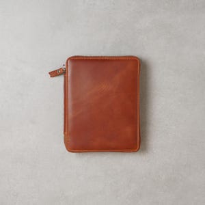 May include: A rectangular, cognac-colored leather zip-around folio or notebook cover. The exterior features a smooth, polished finish with visible natural grain variations and a matching leather zipper pull attached to a silver-toned metal zipper.