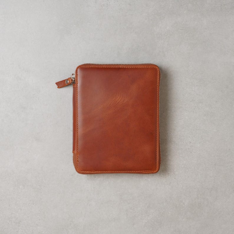 May include: A rectangular, cognac-colored leather zip-around folio or notebook cover. The exterior features a smooth, polished finish with visible natural grain variations and a matching leather zipper pull attached to a silver-toned metal zipper.