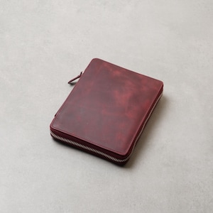 May include: A rectangular, deep burgundy leather portfolio case featuring a smooth, matte finish with subtle tonal variations. The case is secured by a silver-toned metal zipper that wraps around three sides, complete with a matching leather pull tab.