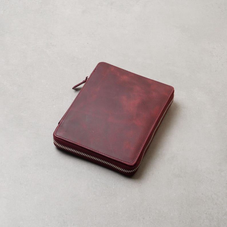 May include: A rectangular, deep burgundy leather portfolio case featuring a smooth, matte finish with subtle tonal variations. The case is secured by a silver-toned metal zipper that wraps around three sides, complete with a matching leather pull tab.