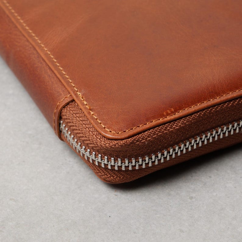 May include: A tan leather portfolio case featuring a smooth, matte surface with visible grain texture. The edges are reinforced with matching tan stitching, and a silver-toned metal zipper runs along the side to secure the closure.