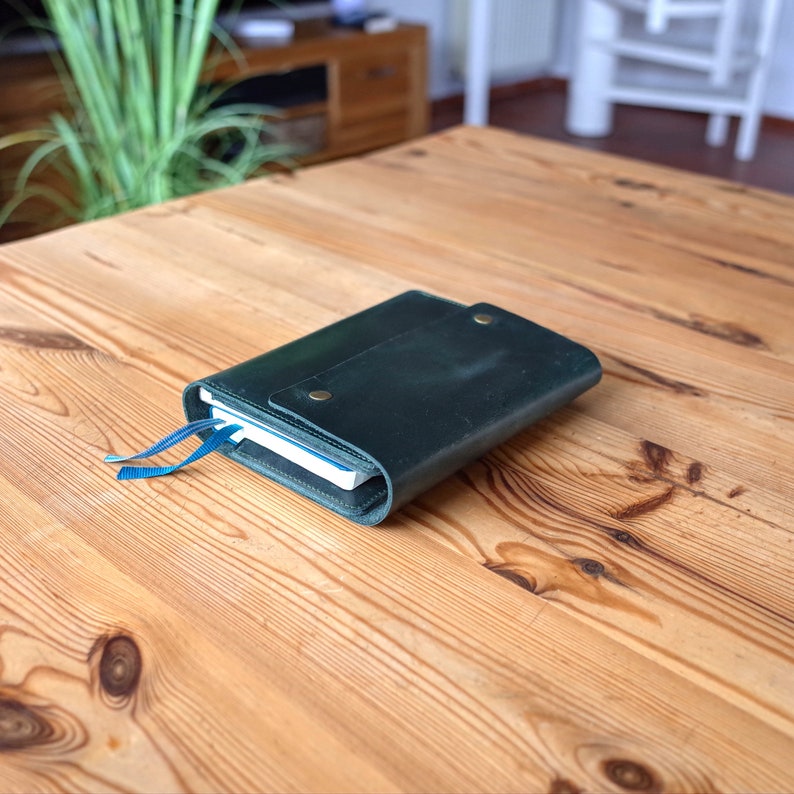 May include: A dark green leather notebook with a blue ribbon bookmark. The notebook is closed and lying on a wooden table.