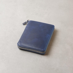 Puede incluir: Una cartera rectangular de cuero azul con un acabado liso y ligeramente jaspeado. La cartera cuenta con una cremallera de metal plateado que recorre tres lados y una pequeña lengüeta de cuero azul a juego en el tirador.