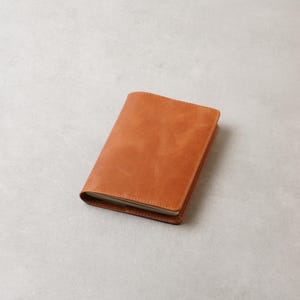 May include: A closed, rectangular, tan leather journal with rounded corners. The journal is lying on a light gray surface. The cover has a smooth, slightly textured appearance.