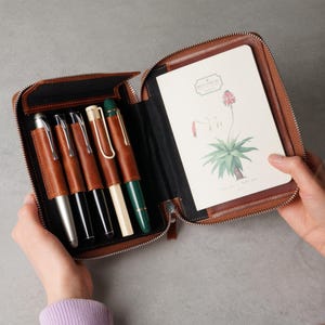May include: A brown leather zippered organizer case containing five pens in individual slots and a notebook with a botanical illustration of a plant and the text Botanical on the cover. The interior features black lining and brown leather accents.
