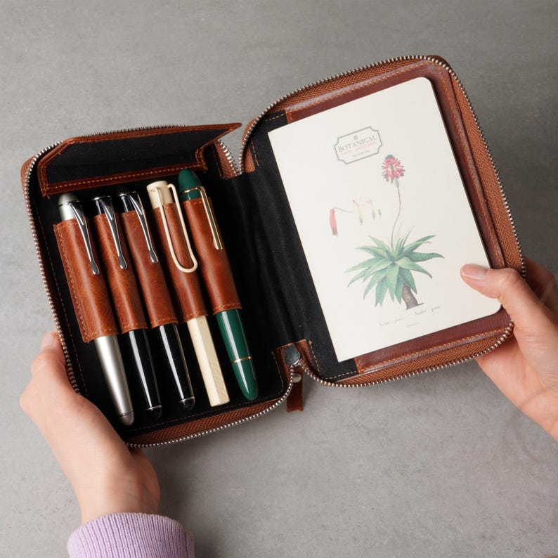 May include: A brown leather zippered organizer case containing five pens in individual slots and a notebook with a botanical illustration of a plant and the text Botanical on the cover. The interior features black lining and brown leather accents.