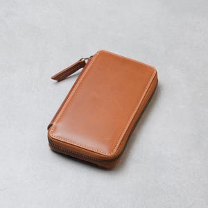 Peut inclure: Un portefeuille rectangulaire en cuir havane avec une finition lisse, des coins arrondis et une fermeture éclair marron foncé qui s'étend sur trois côtés. Une tirette en cuir assorti est fixée au curseur métallique en haut.