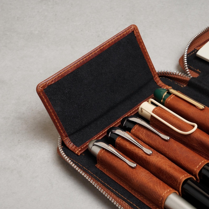 May include: A brown leather zippered pen case featuring a black interior lining with individual slots for writing instruments. The case holds several pens with silver, black, and cream-colored clips, secured within the structured leather sleeves.