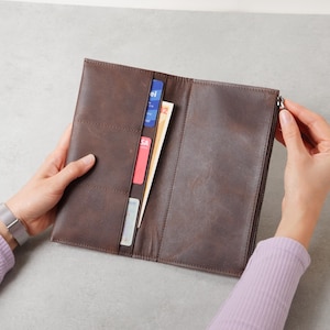 May include: A dark brown leather long wallet featuring multiple vertical card slots on the left side, a central compartment containing paper currency, and a zippered section on the right side, all constructed with visible stitching.