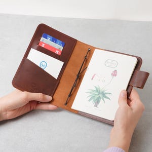 May include: A brown leather notebook cover with an open design, featuring interior card slots holding several cards and a white business card with a blue lightbulb icon. A botanical notebook with a plant illustration is secured by black elastic.