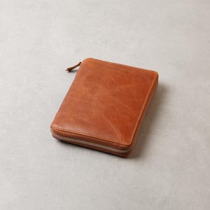 May include: A rectangular, tan-coloured zippered case with a visible grain texture. The case features a silver-toned metal zip that runs along three sides, meeting at a small pull tab located at the top corner of the structured, flat-surfaced item.