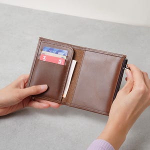 May include: A brown leather wallet featuring a vertical card slot containing two cards, one blue and one red, and a side compartment with a silver metal zip closure along the right edge. The interior shows a smooth finish and visible stitching.