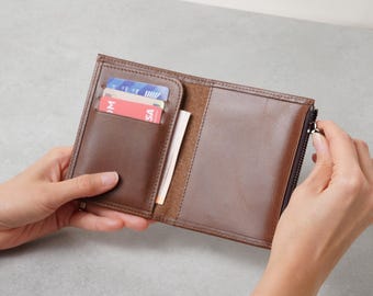 May include: A brown leather wallet featuring a vertical card slot containing two cards, one blue and one red, and a side compartment with a silver metal zipper closure along the right edge. The interior shows a smooth finish and visible stitching.