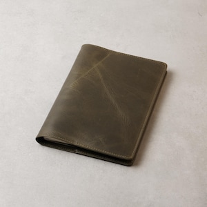 Peut inclure: Un journal ou carnet en cuir vert olive ferm&eacute;. La couverture pr&eacute;sente un aspect l&eacute;g&egrave;rement vieilli avec des veines visibles et de subtiles variations de couleur. Les bords sont soigneusement cousus, pour un design simple et classique.