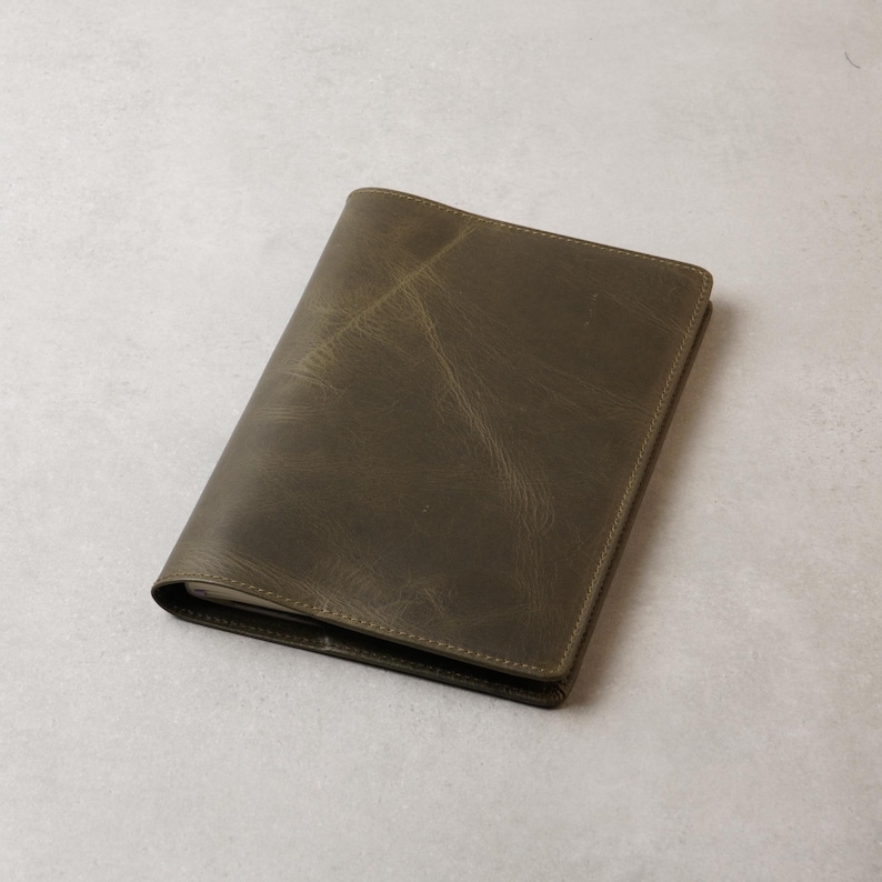 Peut inclure: Un journal ou carnet en cuir vert olive ferm&eacute;. La couverture pr&eacute;sente un aspect l&eacute;g&egrave;rement vieilli avec des veines visibles et de subtiles variations de couleur. Les bords sont soigneusement cousus, pour un design simple et classique.