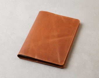 May include: A rectangular notebook cover crafted from smooth, tan-coloured leather. The surface displays natural grain variations and subtle creases, featuring clean, straight stitched edges along the perimeter of the folded leather casing.