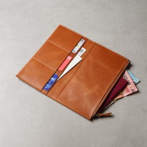 May include: A tan leather long wallet featuring multiple interior card slots, a vertical slip pocket, and a zippered compartment. The wallet is open, revealing several credit cards, a white paper insert, and paper currency tucked inside.