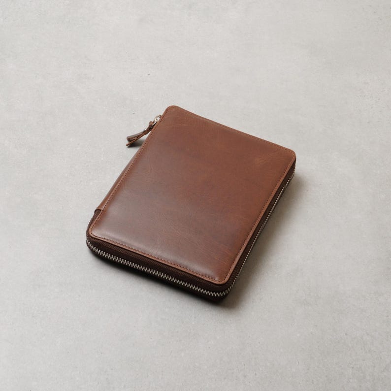 May include: A rectangular brown leather zippered case with a smooth finish and a matching leather zipper pull. The case features a silver-toned metal zipper that runs along three sides, providing a secure closure for the contents inside.