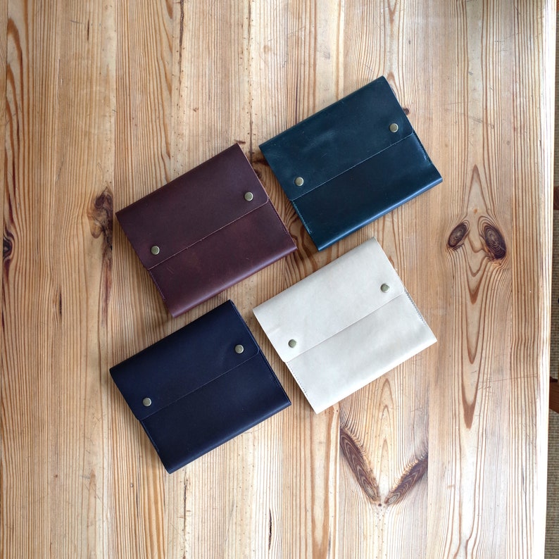 May include: Four leather pouches in different colors: brown, black, beige, and dark blue. Each pouch has a flap closure with a brass button.