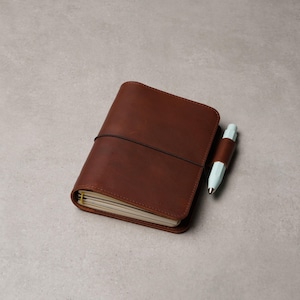 May include: A brown leather journal with a black elastic closure and a light blue pen with a brown leather band. The journal is closed and has a stack of white pages. The pen is resting next to the journal.