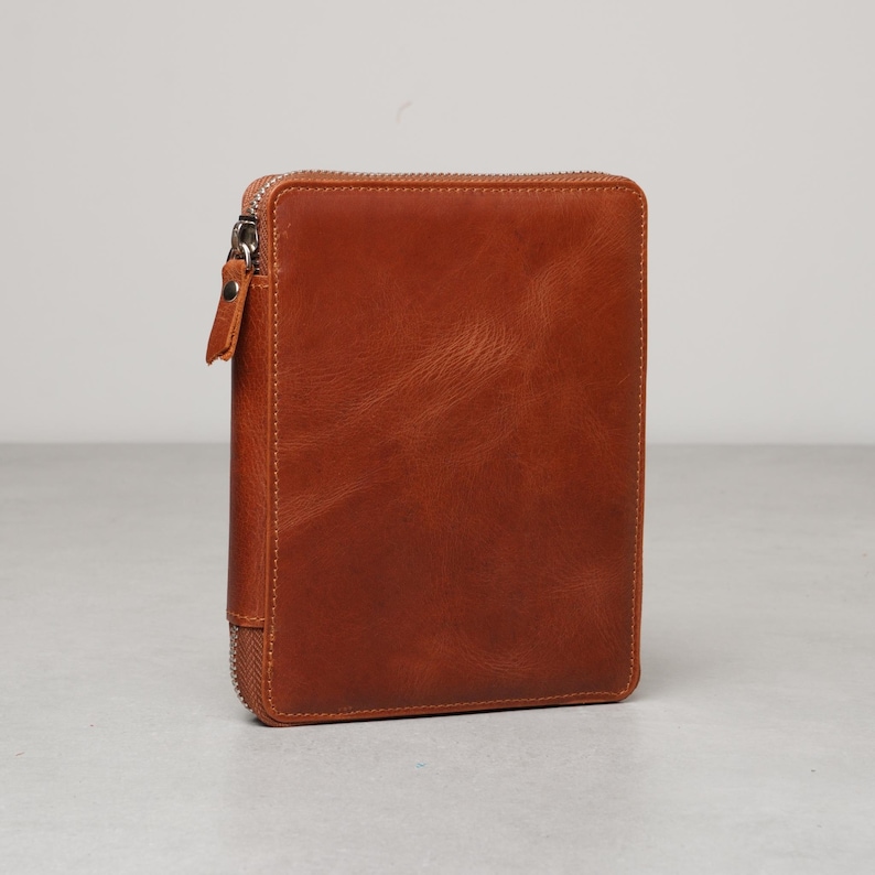 May include: A rectangular, tan-colored leather case with a smooth, slightly textured surface. The case features a silver-toned metal zipper that runs along the left side and top edge, complete with a matching leather pull tab attached to the zipper.