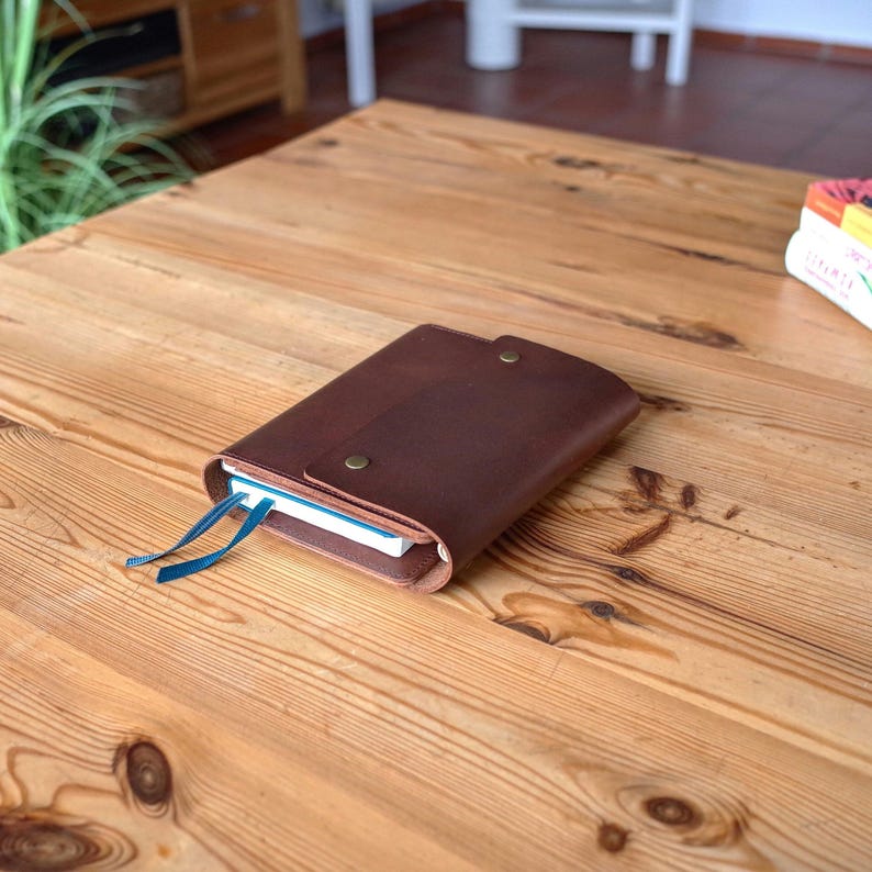 May include: A brown leather notebook cover with a flap closure and two blue ribbon bookmarks. The cover has two brass rivets on the front.
