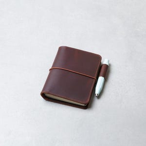 Op de afbeelding: Een donkerbruine lederen notitieboekhoes met afgeronde hoeken, vastgezet met een dun elastiek. Een lichtblauwe pen wordt op zijn plaats gehouden door een bijpassende lederen penlus die aan de zijkant van de hoes is bevestigd.