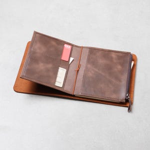 May include: A brown leather notebook cover featuring an open interior with multiple card slots, a central elastic cord for securing paper inserts, and a side zipper pocket. The interior displays a red card and a white card tucked into slots.