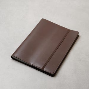 May include: A brown leather journal with a matching elastic band closure. The journal has a smooth, matte finish and a stitched border. The cover is a solid, warm brown color.