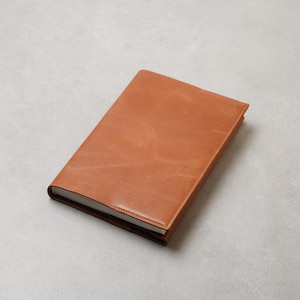 Può includere: Un taccuino rettangolare caratterizzato da una copertina liscia color marrone chiaro con visibili texture a grana naturale. Il taccuino è chiuso e presenta una superficie pulita e uniforme con bordi cuciti lungo il dorso e il perimetro.