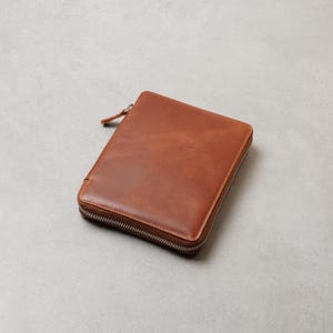 May include: A rectangular, tan-colored leather zippered case with a smooth, polished finish. The case features a silver-toned metal zipper that runs along three sides, with a matching leather pull tab attached to the zipper slider at the top corner.