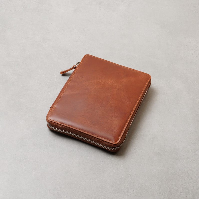 May include: A rectangular, tan-colored leather zippered case with a smooth, polished finish. The case features a silver-toned metal zipper that runs along three sides, with a matching leather pull tab attached to the zipper slider at the top corner.