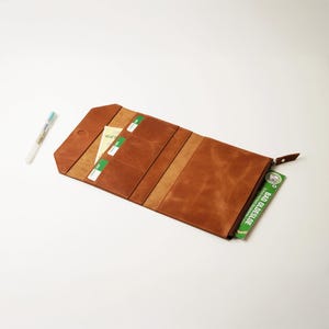 May include: A brown leather wallet, open to display multiple card slots and a zippered compartment. The wallet is rectangular with a snap closure and a brown leather zipper pull. A pen is visible to the left of the wallet.