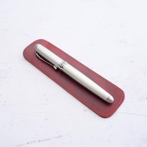 May include: A silver fountain pen with a chrome clip and accents rests on a burgundy leather pen sleeve. The pen sleeve is rectangular with rounded edges and red stitching.