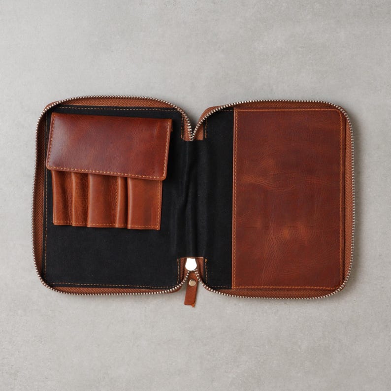 May include: A brown leather zippered organizer case featuring a black interior lining. The left side includes a flap pocket with four vertical slots for pens or tools, while the right side features a large, flat rectangular pocket for documents.