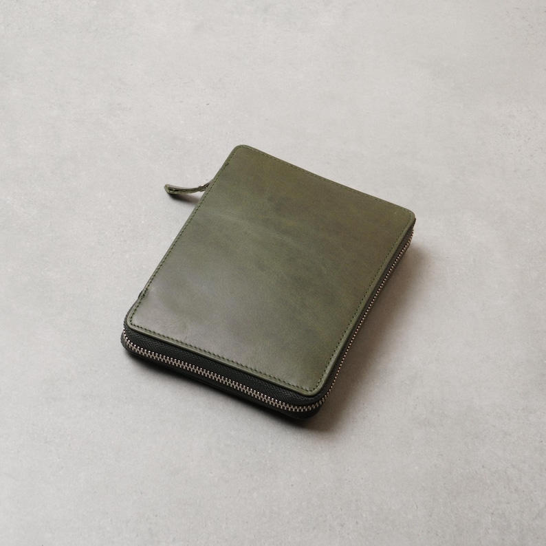 May include: A rectangular olive green leather case with a smooth, matte finish and rounded corners. The case features a silver-toned metal zipper that runs along three sides, complete with a matching leather pull tab attached to the zipper slider.