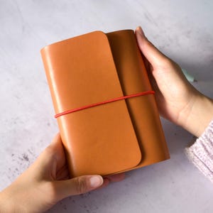 Puede incluir: Un diario de cuero marrón claro sostenido por dos manos. El diario está asegurado con una banda elástica roja. La cubierta está doblada y las páginas son visibles. El diario tiene un tono cálido y terroso.