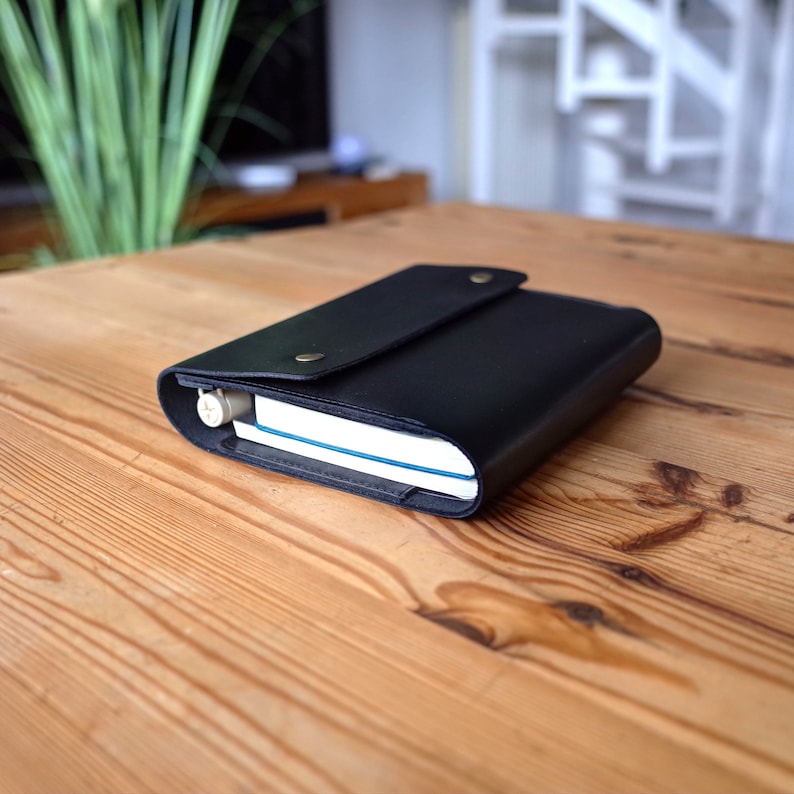 May include: A black leather notebook cover with a flap closure and a small metal button. The cover is closed and a notebook is visible inside.