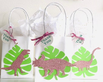 May include: Three white gift bags with green and pink dinosaur designs. Each bag has a pink and green leaf with a pink glitter dinosaur. The bags have white tissue paper inside and a pink ribbon with a round tag that says "Thank You".