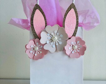 May include: White gift bag with pink and gold bunny ears and pink paper flowers. The bag has white handles.
