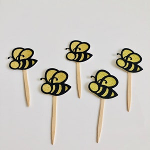 May include: Five bee-shaped cupcake toppers. Each topper features a black and gold glitter bee design attached to a wooden stick. The bees have black stripes and gold glitter wings.