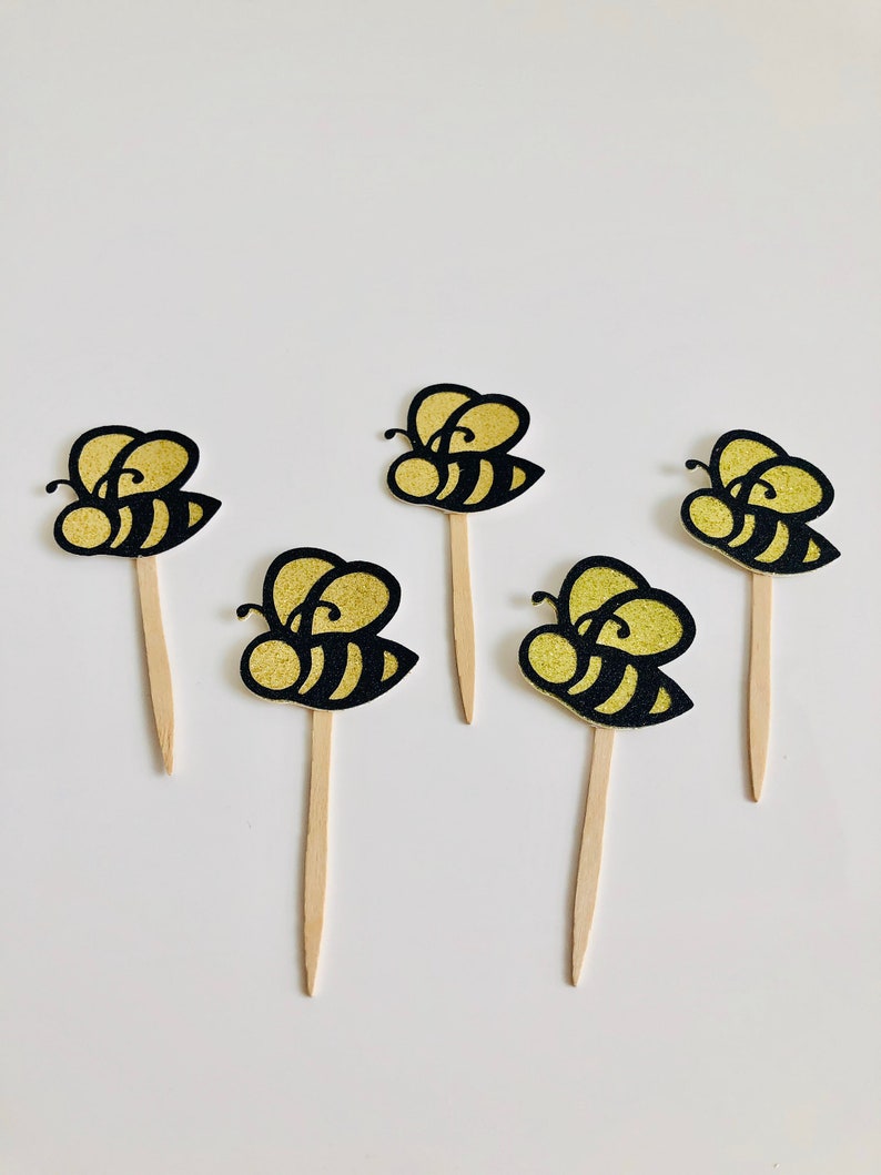 May include: Five bee-shaped cupcake toppers. Each topper features a black and gold glitter bee design attached to a wooden stick. The bees have black stripes and gold glitter wings.