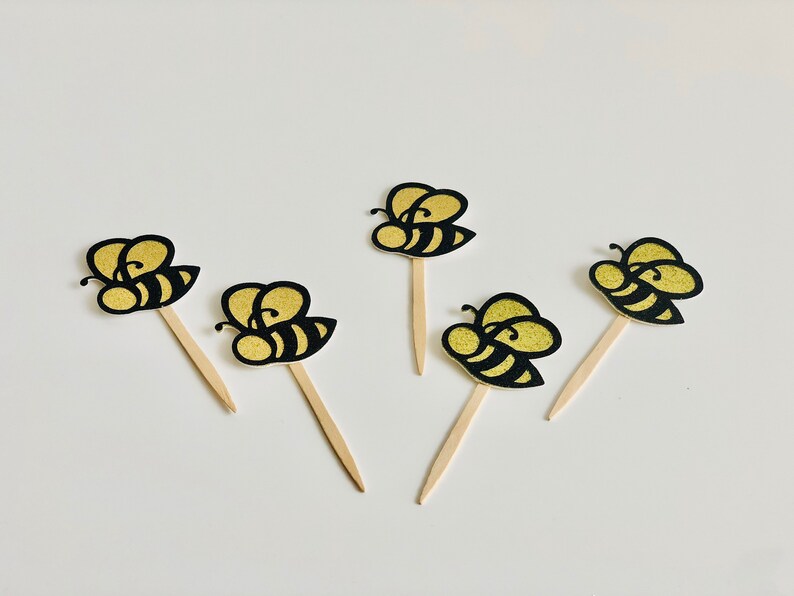 May include: Five decorative bee-shaped cupcake toppers. Each bee has a gold glitter body with black stripes and a wooden stick. The toppers are arranged on a white surface.